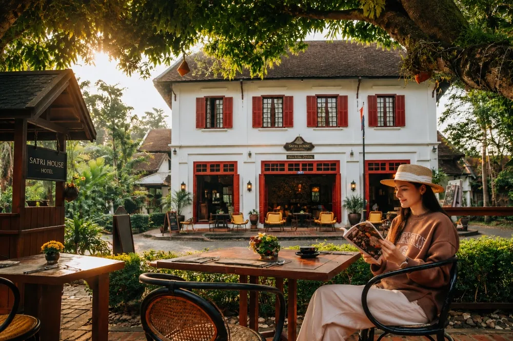 A woman reading a book at a cafe terrace in front of a colonial-style hotel